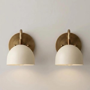 May include: A pair of wall sconces featuring a circular brushed brass backplate and a curved arm supporting a matte off-white dome-shaped shade. The design includes a small brass finial detail on each side of the arm where it meets the backplate.