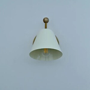 May include: A white, cone-shaped wall sconce with a brass-coloured base and a small, round finial. The light fixture has a visible bulb and is mounted on a light blue wall. The sconce is a modern lighting fixture.