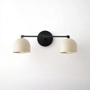 May include: A modern wall-mounted light fixture with two cream-colored, dome-shaped shades. The shades are attached to a black horizontal bar and a circular black base. The fixture is designed for indoor use and provides ambient lighting.