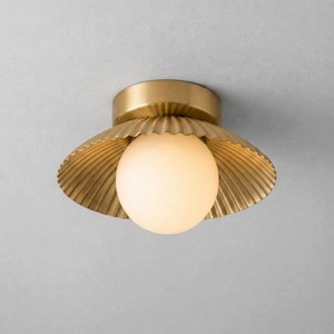 May include: A flush mount ceiling light fixture featuring a circular brass base and a wide, scalloped, metallic gold shade. A single, spherical, frosted white glass globe is positioned in the center, emitting a soft, warm, diffused light.