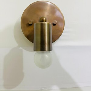 May include: A wall-mounted light fitting featuring a circular brass backplate with a brushed finish. A cylindrical brass socket extends from the centre, holding a single, small, round, frosted white glass light bulb. Two small screws are visible.