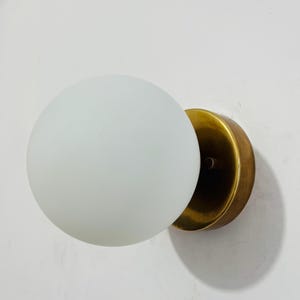 May include: A wall-mounted light fixture featuring a spherical, frosted white glass globe shade attached to a circular, brushed brass-toned metal base. The design consists of a simple, clean silhouette with a metallic mounting plate and a glass orb.