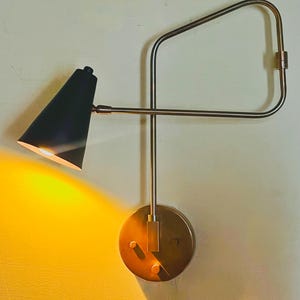 May include: A wall-mounted lamp featuring a circular brass base and a slender, adjustable brass arm. The lamp has a conical black shade that emits a warm, bright yellow light, illuminating the area to the left of the fixture with a soft glow.