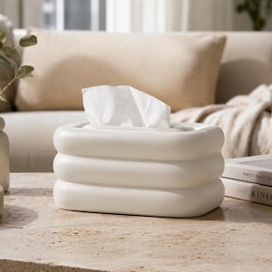 May include: A rectangular off-white tissue box cover featuring a rounded, three-tiered stacked design. The smooth, matte-finished object has a central opening at the top from which a single white facial tissue is pulled upward and partially exposed.