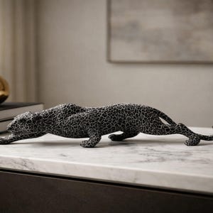May include: A black and white leopard ornament with a textured design, shown crawling. The ornament is on a white marble surface. A gold sphere and books are in the background. The leopard is a decorative item.