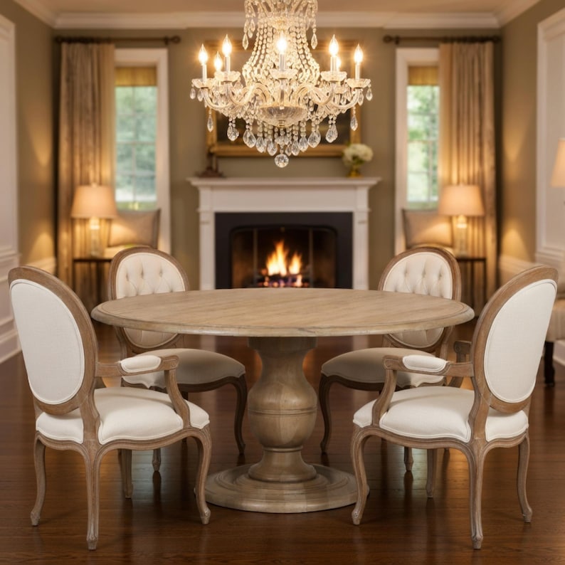 May include: A dining room set featuring a round wooden table and four upholstered chairs. The chairs have light-colored fabric and wooden frames. A crystal chandelier hangs above the table, and a fireplace is in the background.