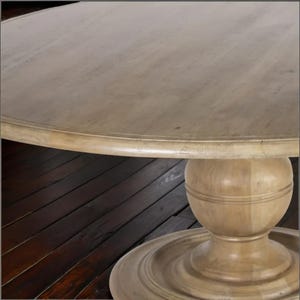 May include: A round wooden table with a light-colored, distressed finish. The table features a pedestal base with a bulbous center and a circular foot. The tabletop has a subtle, aged appearance, and the overall design is rustic.