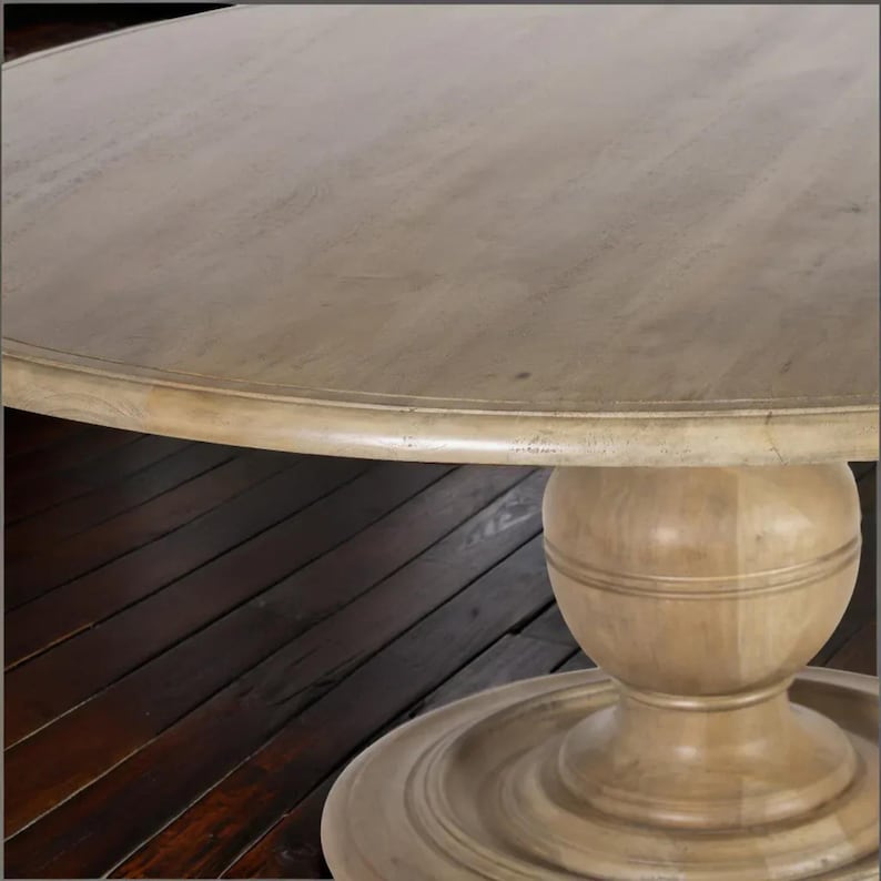 May include: A round wooden table with a light-colored, distressed finish. The table features a pedestal base with a bulbous center and a circular foot. The tabletop has a subtle, aged appearance, and the overall design is rustic.