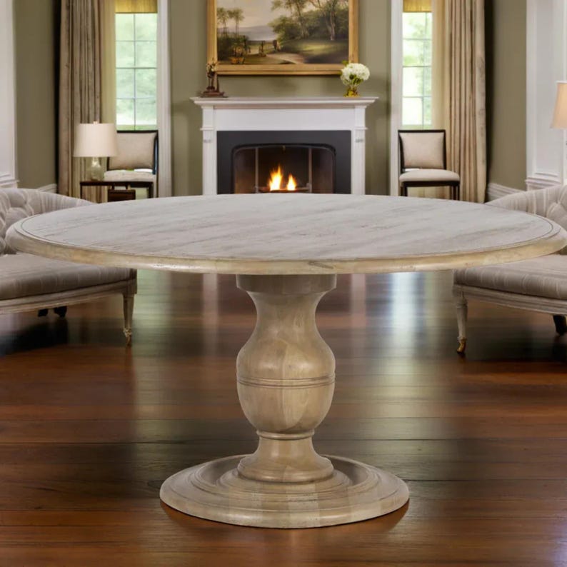 May include: A round, light-colored wooden dining table with a pedestal base. The tabletop has a subtle wood grain pattern. The table is set in a well-lit room with a fireplace and chairs in the background. The table is approximately 60 inches in diameter.