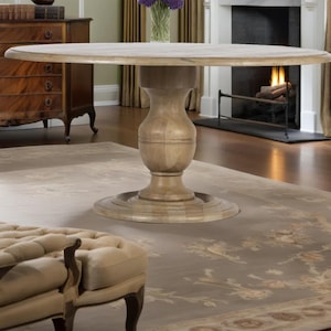 May include: A light-colored, round wooden dining table with a pedestal base. The table is set in a room with a fireplace, a patterned rug, and a tufted ottoman. The table's diameter is approximately 60 inches.