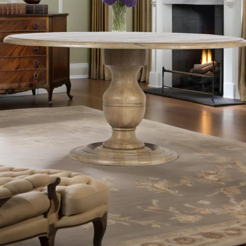 May include: A light-colored, round wooden dining table with a pedestal base. The table is set in a room with a fireplace, a patterned rug, and a tufted ottoman. The table's diameter is approximately 60 inches.