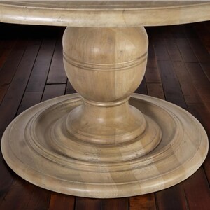 May include: A light-colored wooden pedestal table base. The base has a round, tiered design with a central, bulbous support. The wood grain is visible, and the finish has a slightly distressed appearance. The table appears to be sturdy and well-crafted.