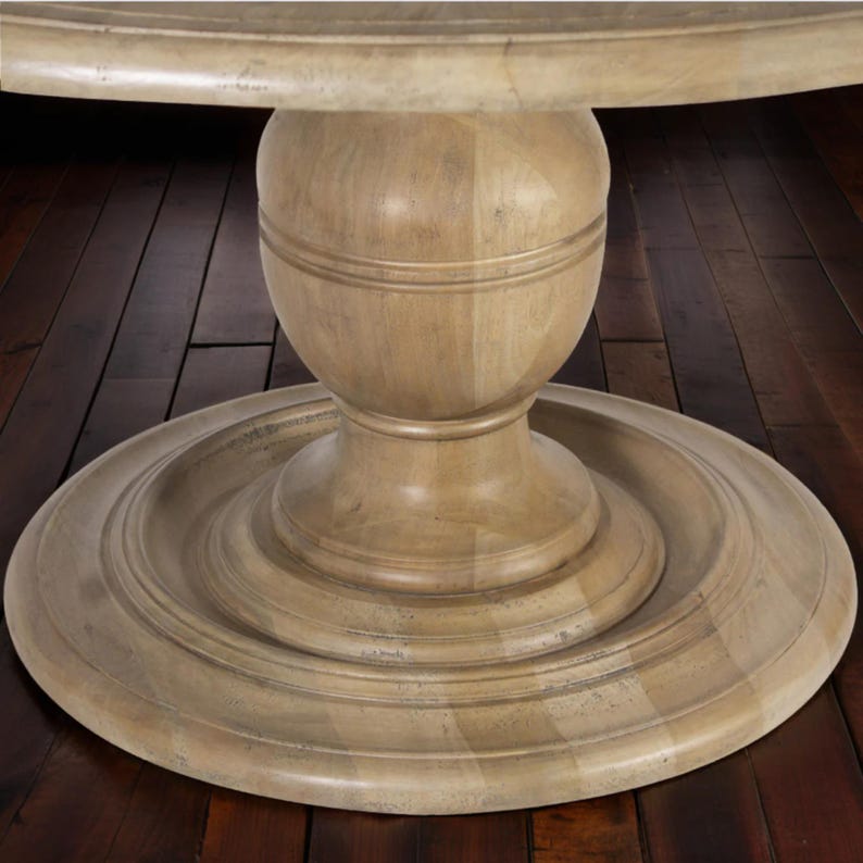 May include: A light-colored wooden pedestal table base. The base has a round, tiered design with a central, bulbous support. The wood grain is visible, and the finish has a slightly distressed appearance. The table appears to be sturdy and well-crafted.