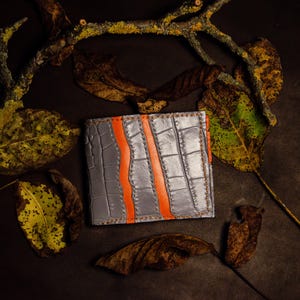 May include: A gray leather wallet with an orange stripe design. The wallet has a textured, crocodile-like pattern. The image is set against a dark background with dried leaves and a branch.