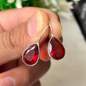 May include: Teardrop earrings featuring a deep red gemstone set in silver-coloured metal. The earrings hang from a simple hook, highlighting the polished, faceted stones. The gemstones have a rich, deep colour, creating a striking contrast.