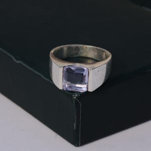 May include: A silver ring with a rectangular, light purple gemstone. The ring has a thick band and a simple, classic design. The gemstone is set in a square bezel, creating a bold and elegant look. The ring is displayed on a dark surface.