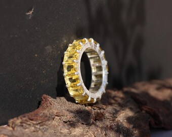 May include: A silver ring with a band of rectangular, yellow gemstones. The ring is set against a dark background and rests on a piece of wood. The gemstones are evenly spaced around the band.