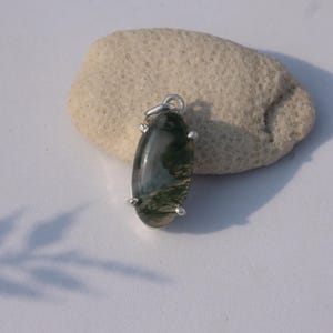 Puede incluir: Un colgante ovalado con una argolla plateada y cuatro garras plateadas. El colgante presenta una piedra de ágata musgosa con patrones verdes y blancos. El colgante se muestra sobre una roca clara.