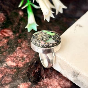 Peut inclure: Une bague en argent avec une grosse pierre ronde et mouchetée. La pierre présente un mélange de teintes noires, vertes et roses. La bague est posée sur une surface blanche, avec des fleurs blanches en arrière-plan. Un bijou unique.