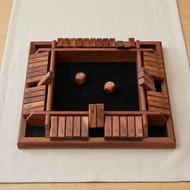 May include: A wooden Shut the Box game with a dark felt playing surface. The game features numbered wooden tiles and two dice. The frame is made of dark wood and is set on a neutral-colored surface.
