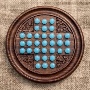 May include: A wooden solitaire board game with a circular design. The board is made of dark brown wood and features carved floral patterns. Thirty-three light blue marbles are arranged in the game's starting position.