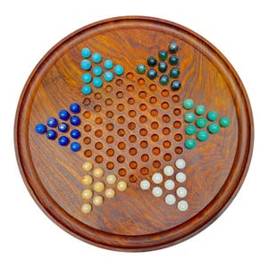 May include: A wooden Chinese Checkers board with a star-shaped pattern of holes. The board is filled with colorful marbles in shades of blue, green, yellow, and white. The board is round and made of a dark wood with a visible wood grain.