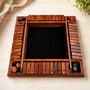 May include: A wooden Shut the Box dice game with a dark interior and numbered wooden tiles. The game includes several dice and is set on a light-colored surface. The numbers 1 through 12 are visible on the tiles.