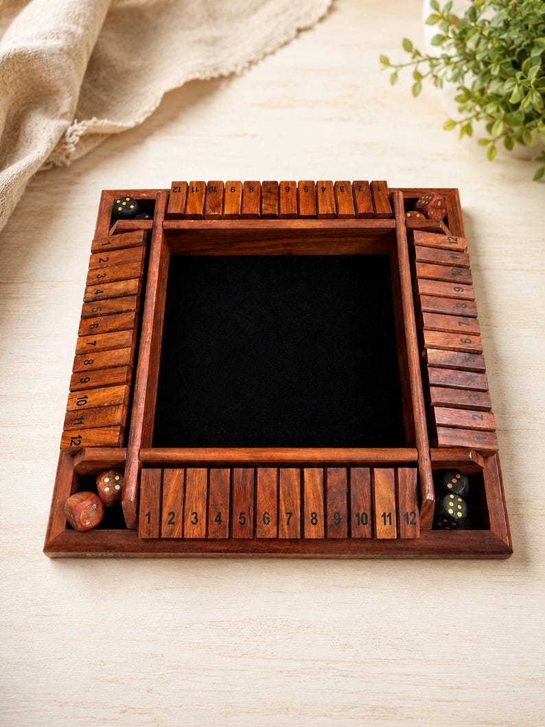 May include: A wooden Shut the Box dice game with a dark interior and numbered wooden tiles. The game includes several dice and is set on a light-colored surface. The numbers 1 through 12 are visible on the tiles.