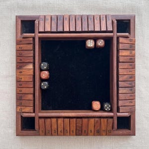 May include: A wooden Shut the Box dice game with a dark wood finish. The game board has a black felt center and numbered wooden tiles. Several dice are scattered on the board, ready to be rolled. The numbers 1-12 are printed on the sides.