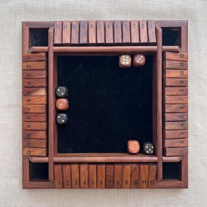 May include: A wooden Shut the Box dice game with a dark wood finish. The game board has a black felt center and numbered wooden tiles. Several dice are scattered on the board, ready to be rolled. The numbers 1-12 are printed on the sides.