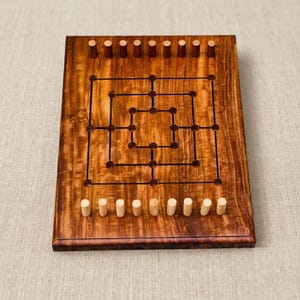 May include: A wooden Nine Men's Morris board game. The board is a rich brown color with a visible wood grain. It features a grid of lines and holes for game pieces. Twelve wooden pegs are included, six light and six dark.
