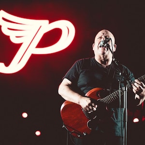 May include: A musician on stage, playing a red electric guitar and singing into a microphone. A large, illuminated white logo with a stylized "P" and wings is in the background. The stage is dark with a few small lights.