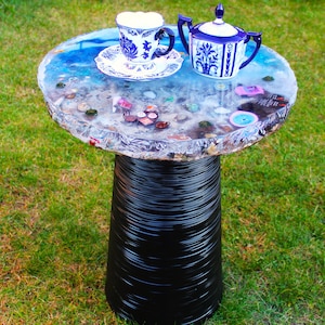 May include: A round side table with a black ribbed pedestal base and a clear resin tabletop featuring embedded miniature objects, including a blue and white porcelain teacup on a saucer and a matching lidded sugar bowl.