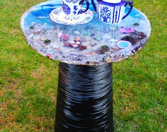 Puede incluir: Una mesa auxiliar redonda con una base de pedestal negra acanalada y un tablero de resina transparente con objetos en miniatura incrustados, incluyendo una taza de té de porcelana azul y blanca y un azucarero a juego.