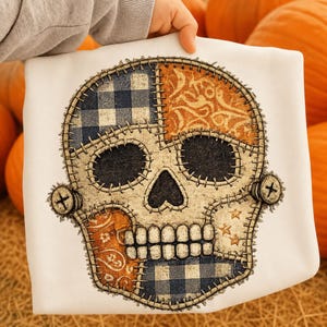 Può includere: Una t-shirt bianca con una grande grafica di un teschio composto da vari ritagli di tessuto. Il design del teschio include sezioni a quadretti blu e bianchi, motivi paisley arancioni e crema, il tutto delineato da cuciture scure e bulloni.