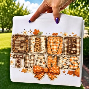 Op de afbeelding: Een wit sweatshirt met de tekst GIVE THANKS in grote blokletters. Elke letter heeft een uniek patroon, waaronder bruine ruitjes, luipaardprint en oranje gingham. Het ontwerp bevat strikken, herfstbladeren en kleine bloemen.