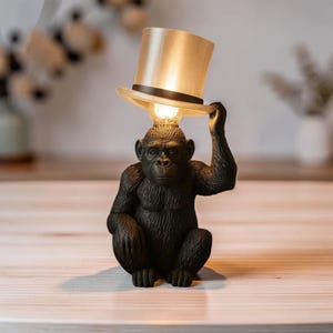 May include: A whimsical table lamp featuring a seated gorilla figure holding a top hat that serves as a lampshade. The lamp has a warm, soft glow, and the gorilla is a dark, textured color.