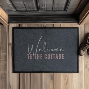 Könnte beinhalten: Eine rechteckige graue Fußmatte mit schwarzem Rand, vor einer Holztür platziert. Auf der Matte steht in weißer und rosa Schrift "Welcome to the Cottage". Braune Lederschuhe sind rechts zu sehen.