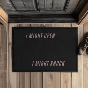 Könnte beinhalten: Schwarze rechteckige Fußmatte mit dem Text "I MIGHT OPEN" über "I MIGHT KNOCK" in hellrosa Schrift. Die Matte liegt auf einem Holzboden, mit einem Paar brauner Schuhe rechts.