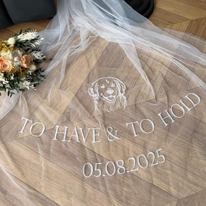 May include: A delicate white wedding veil featuring embroidered text: "TO HAVE & TO HOLD" and the date "05.08.2025". The veil has an embroidered dog design. A bouquet of flowers is visible in the background.