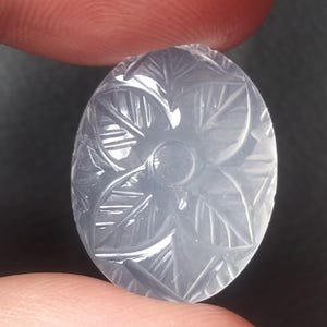 May include: Oval-shaped, translucent gemstone with a carved floral design. The light gray stone features a central flower with surrounding leaves, all intricately carved. The gemstone is held between two fingers.