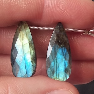 May include: Two faceted labradorite teardrop-shaped gemstones. The stones display a mix of blue, green, and gray hues, with a shimmering, iridescent effect. The gemstones are strung on a thin, silver-colored wire, ready for jewelry making.