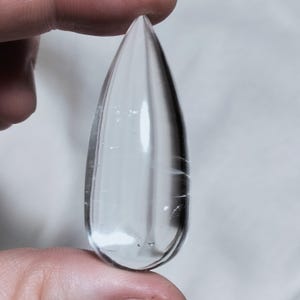 May include: A clear, teardrop-shaped crystal pendant. The polished gemstone is held between fingers, showcasing its transparency and internal striations. The crystal's pointed end and smooth surface reflect light, creating a subtle shimmer.