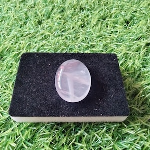 May include: Oval-shaped, translucent rose quartz gemstone with internal veining, resting on a black, rectangular display stand. The gemstone is a pale pink color and is set against a backdrop of green grass.