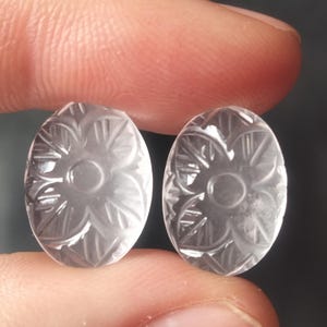 May include: Two clear, oval-shaped gemstones with carved floral designs. The gemstones are held between fingers, showcasing the intricate details of the carved flowers. The gemstones are translucent, allowing light to pass through, highlighting the delicate patterns.