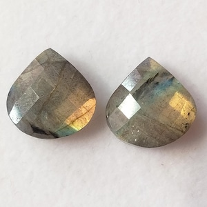 May include: Two pear-shaped labradorite gemstones. The stones are gray with iridescent flashes of blue, yellow, and green. The facets catch the light, creating a sparkling effect. The gemstones are set against a white background.