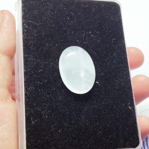 May include: A pale, oval-shaped gemstone, possibly a cabochon, rests on a black velvet surface within a clear plastic case. The stone has a translucent quality, appearing white with subtle internal patterns. The setting is held by a hand.