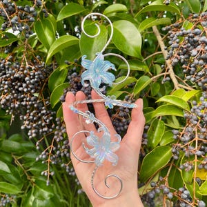 May include: A silver-toned wire ear cuff featuring two iridescent light blue lily-shaped flowers and smaller floral accents. The metal wire is shaped into elegant swirls and curves that wrap around the ear, creating a decorative accessory.
