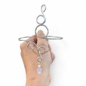 May include: A silver wire arm cuff with an ornamental design. The cuff has a series of loops and swirls, with a small charm of a star-shaped bead and a pearl. The jewellery is held against a white background.