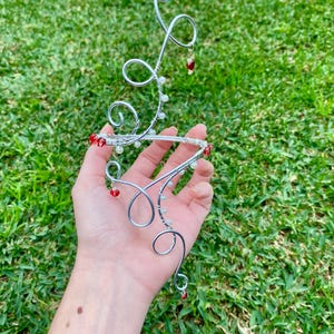 May include: A decorative silver wire piece with a swirling design, decorated with small red and white beads. The piece is held in a hand against a backdrop of green grass.